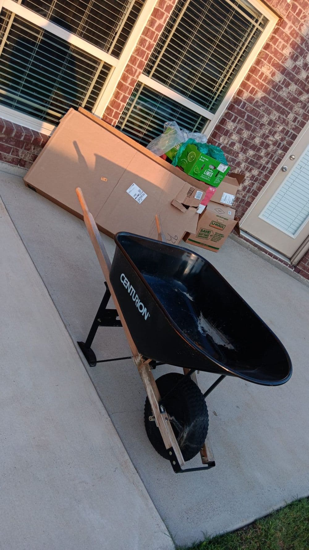 Black wheelbarrow on pavement beside cardboard boxes and bags of soil near a brick wall.