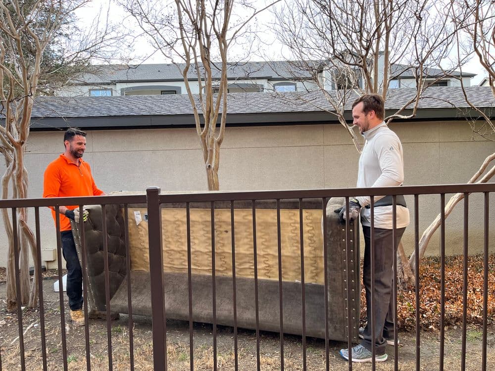 Two men moving a large couch outdoors near a fence and trees.