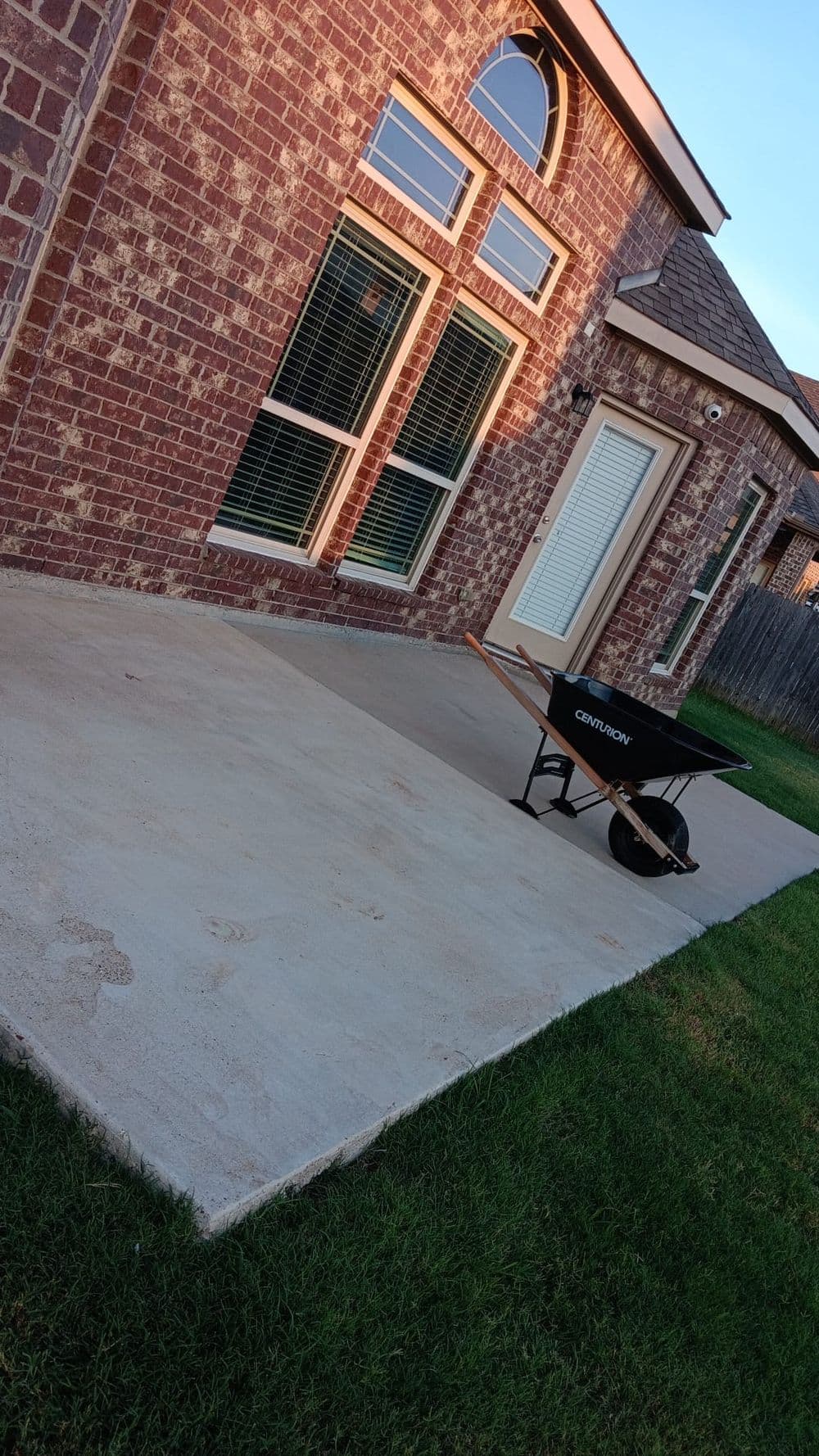 Wheelbarrow on concrete patio next to a brick house with large windows and grass.