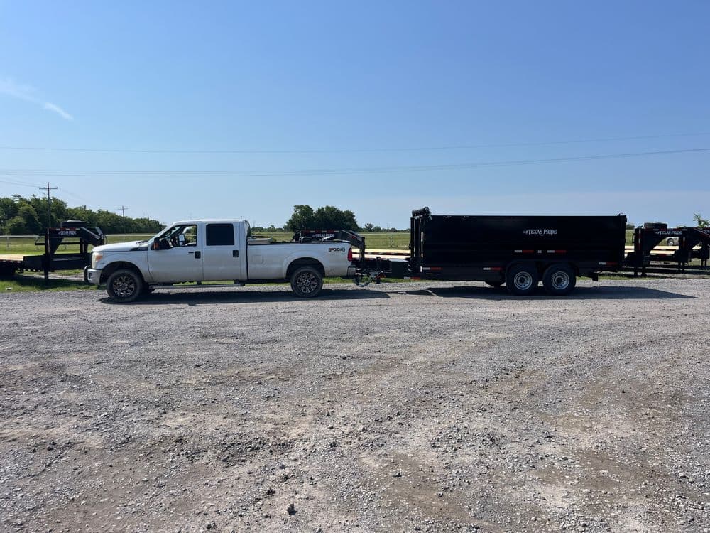 White pickup truck towing a black dump trailer on a gravel lot under a clear blue sky.