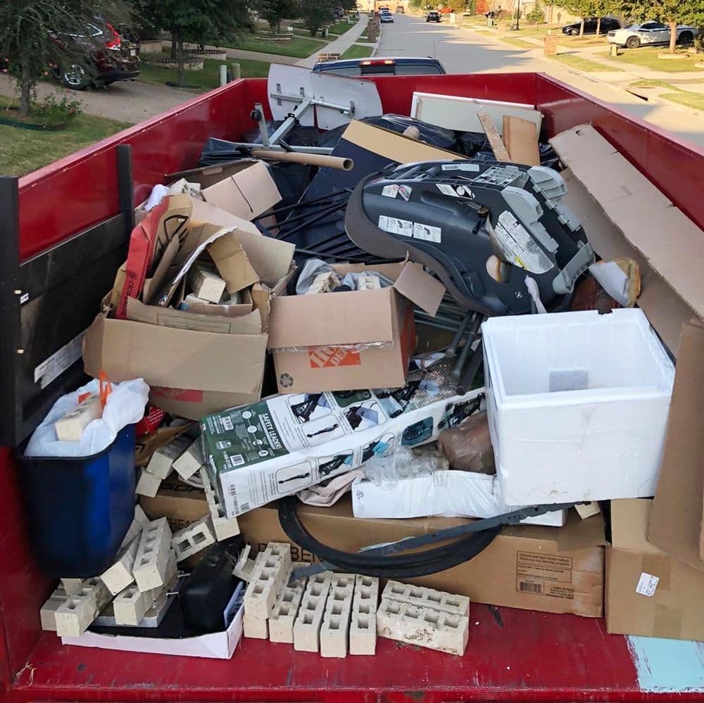 Overflowing dumpster filled with cardboard boxes, furniture parts, and household debris.