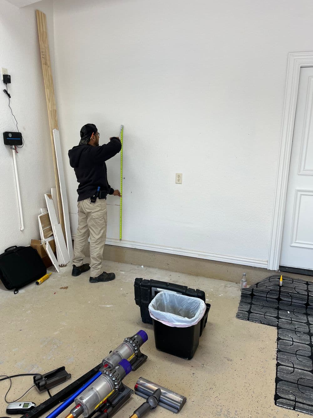 Worker measuring wall in garage with tools and trash bin nearby. Home improvement project.
