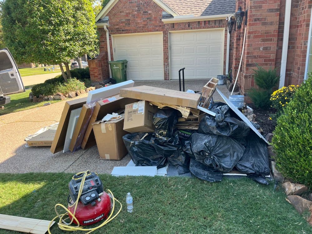 Piled debris and trash bags outside a home, with a lawn and garage in the background.