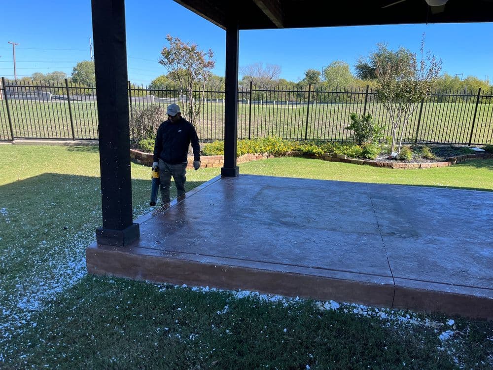 Worker using a leaf blower on a clean patio with a manicured lawn and clear blue sky.