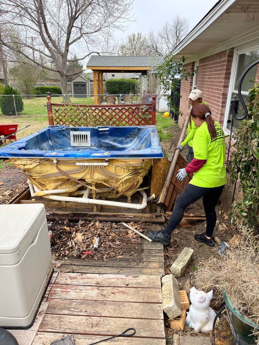 Person cleaning debris around a blue hot tub in a backyard setting, with tools and a white object nearby.