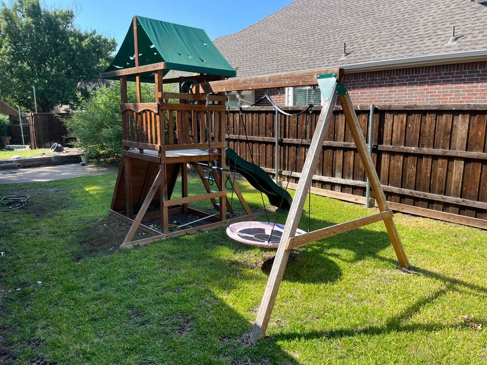 Wooden playset with swings and slide in backyard, surrounded by green grass and wooden fence.